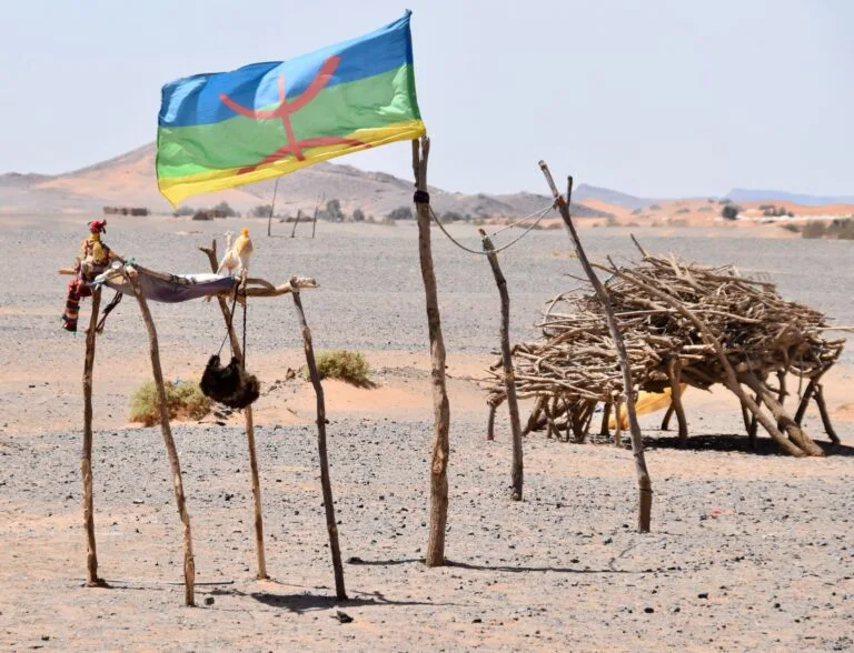 Bandiera berbera blu, rossa e verde con striscia gialla sventola su strutture di legno secco in un paesaggio desertico arido.