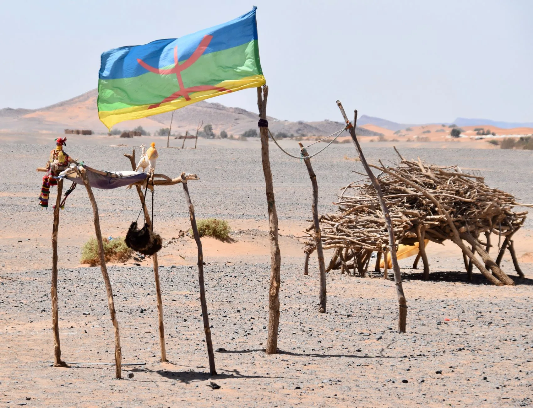 Bandiera berbera blu, rossa e verde con striscia gialla sventola su strutture di legno secco in un paesaggio desertico arido.