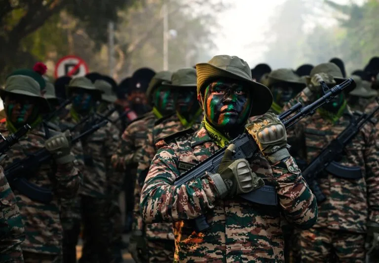 Soldati in uniforme mimetica e volti dipinti con strisce colorate portano fucili automatici in formazione.