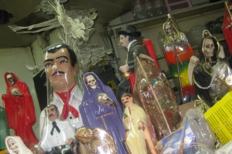 Statuette e figure religiose in plastica e cartone, tra cui raffigurazioni di santi, esposti in uno spazio interno affollato.