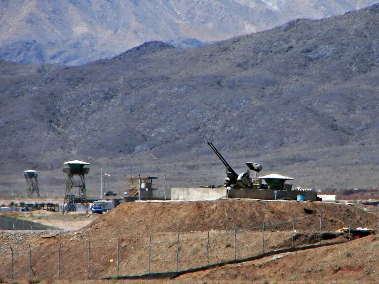 Postazione militare con cannone antiaereo su terrapieno desertico, montagne sullo sfondo e infrastrutture protette da…