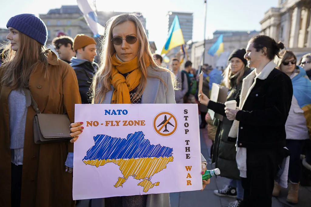 Manifestante con cartello raffigurante l'Ucraina e scritta NATO NO-FLY ZONE tra bandiere blu e gialle.