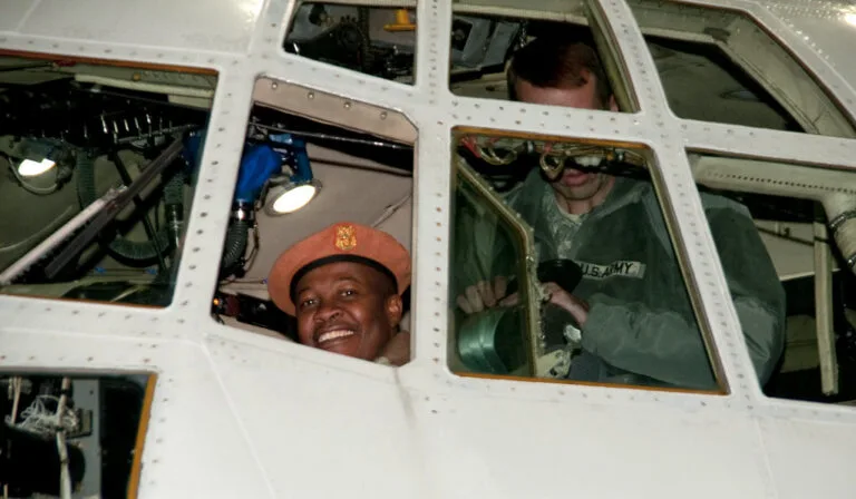 Due persone sorridono dai finestrini di un veicolo militare bianco, una con caschetto arancione.