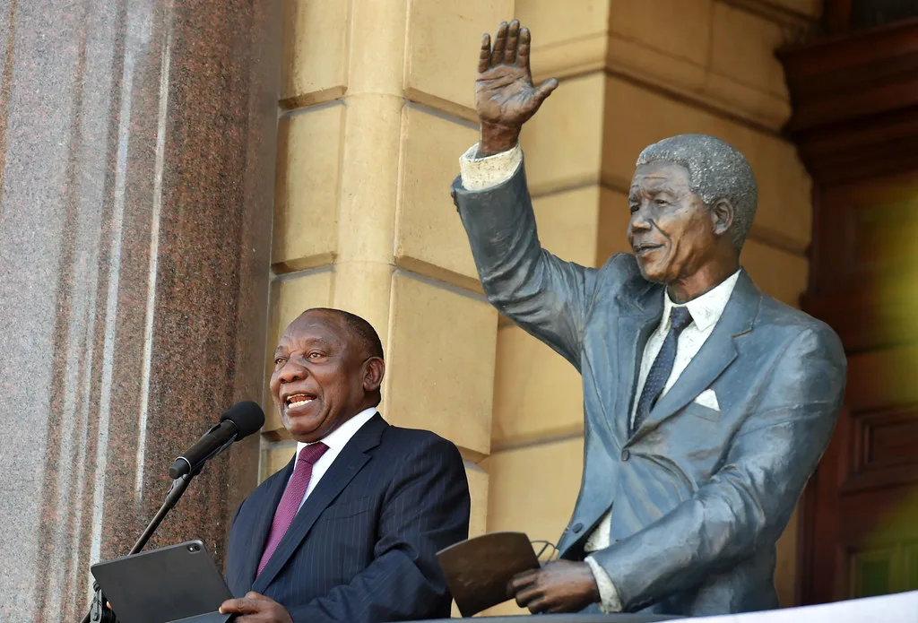 Il presidente sudafricano Cyril Ramaphosa parla al microfono accanto a una statua di Nelson Mandela con il pugno alzato.