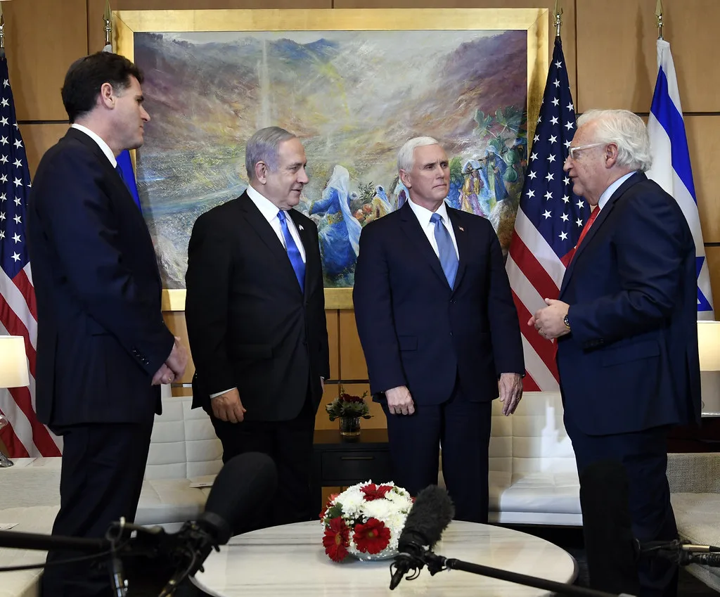 Quattro uomini in abiti scuri conversano in una sala ufficiale con bandiere americane e israeliane alle spalle.