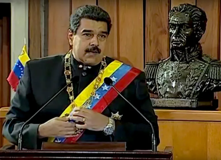 Uomo in abito formale con fascia presidenziale tricolore venezuelana parla al microfono davanti a un busto di bronzo.