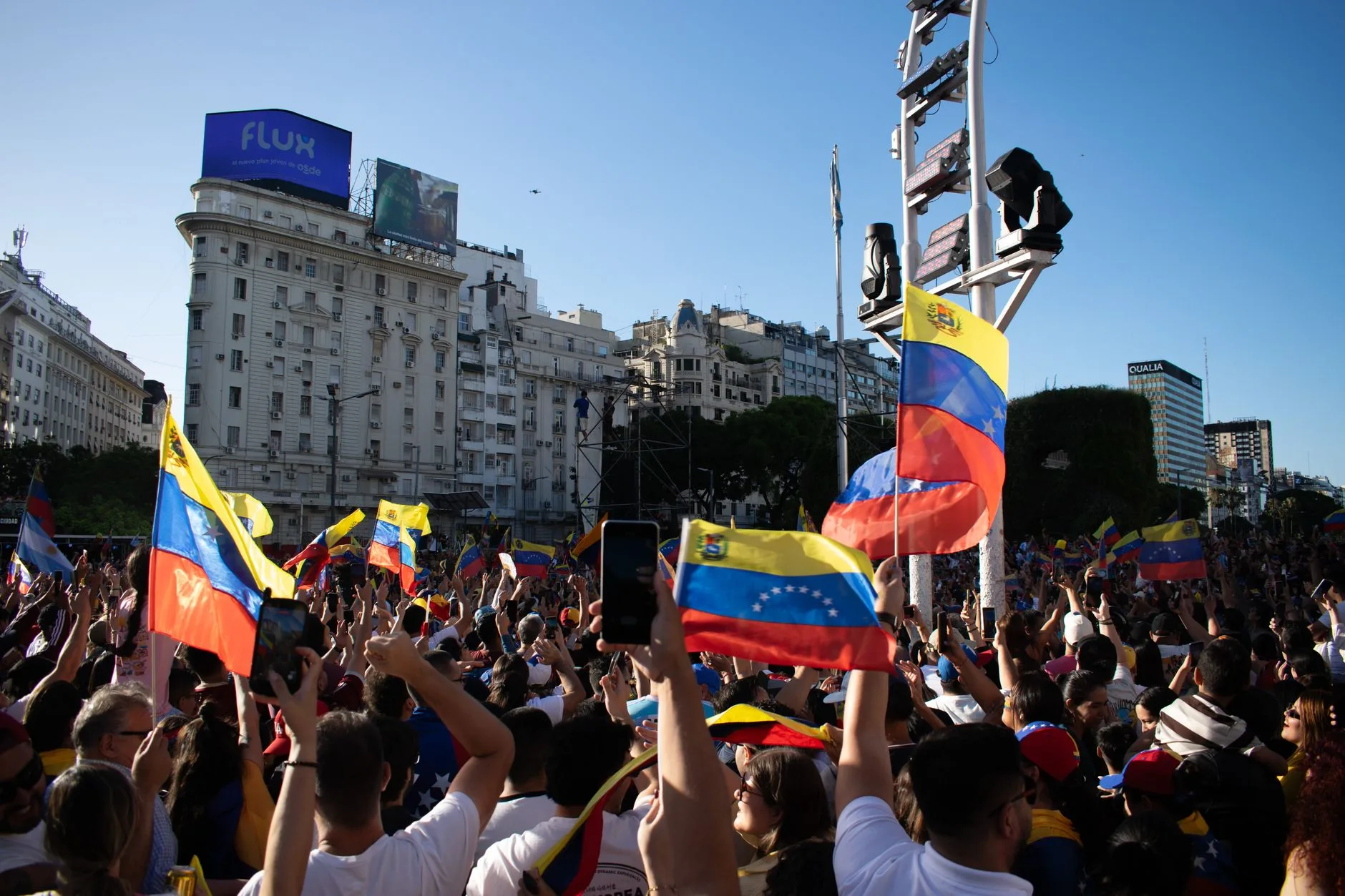 Folla in piazza agita bandiere del Venezuela con i colori giallo, blu e rosso durante una manifestazione di protesta urbana.
