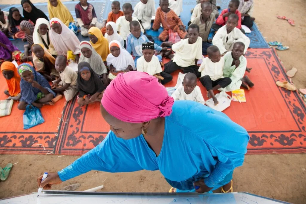 Una donna in abito blu e turbante rosa informa una comunità seduta su tappeti colorati in uno spazio aperto.