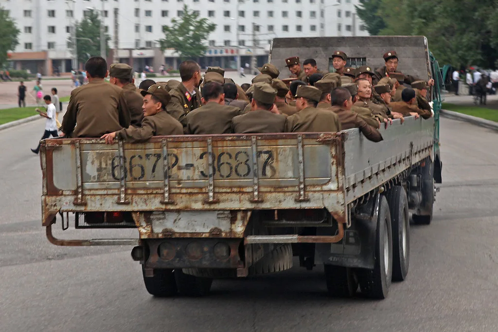 Camion militare carico di soldati in uniforme marrone su strada urbana con edifici residenziali sullo sfondo.