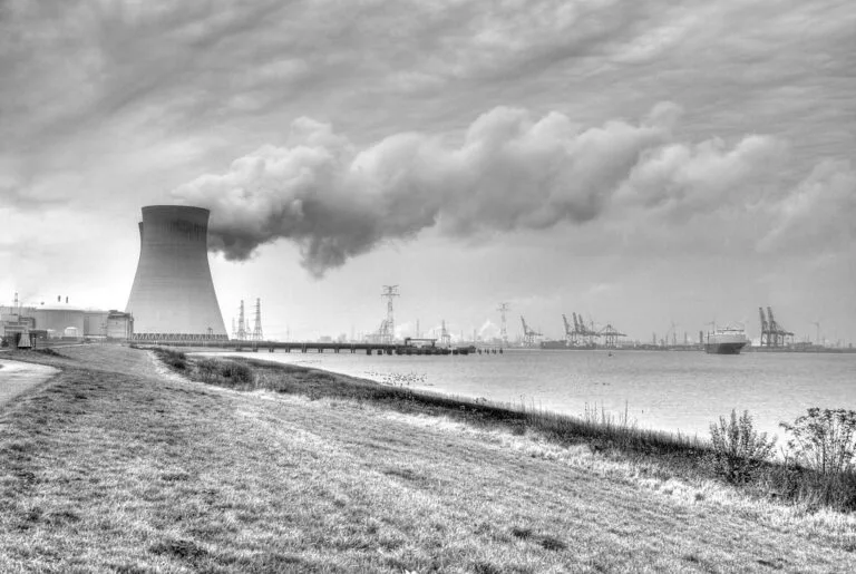 Centrale nucleare con torre di raffreddamento che emette vapore, affacciata su un porto industriale con gru e navi cargo.