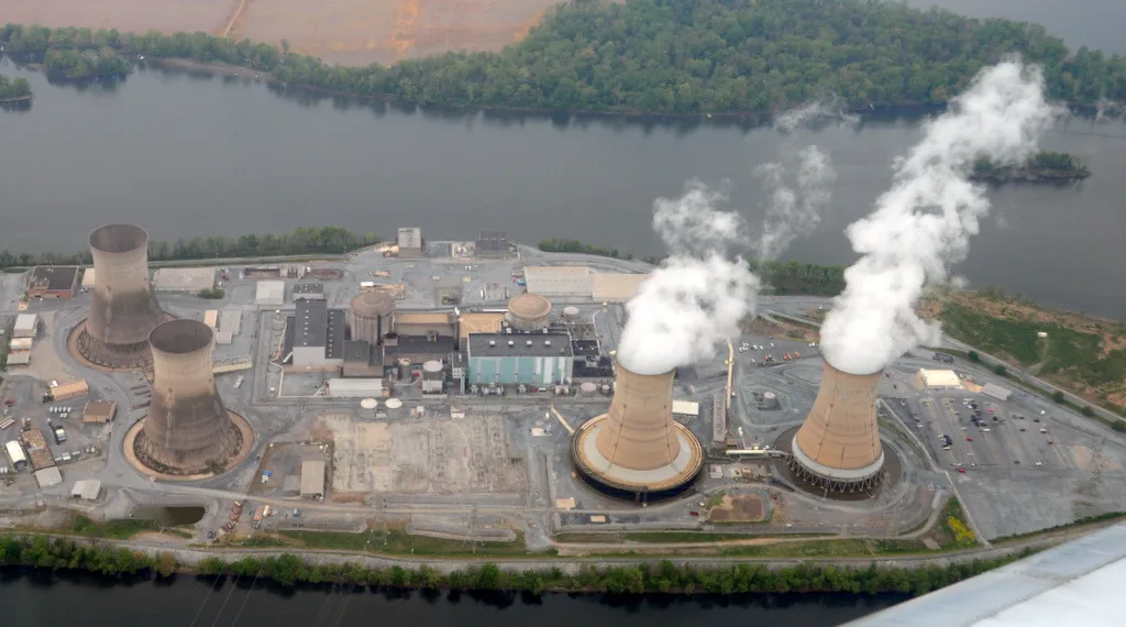 Centrale nucleare vista dall'alto con torri di raffreddamento che emettono vapore, edifici industriali e fiume sullo sfondo.