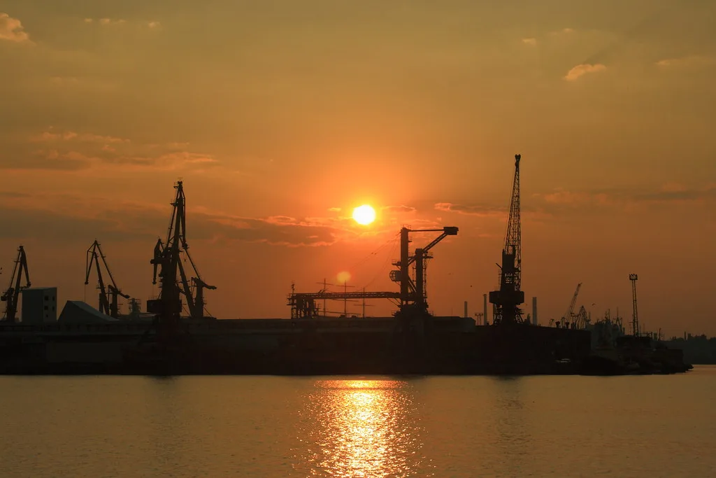 Silhouette di navi cargo e gru portuali al tramonto su acque calme, con sole arancione all'orizzonte.