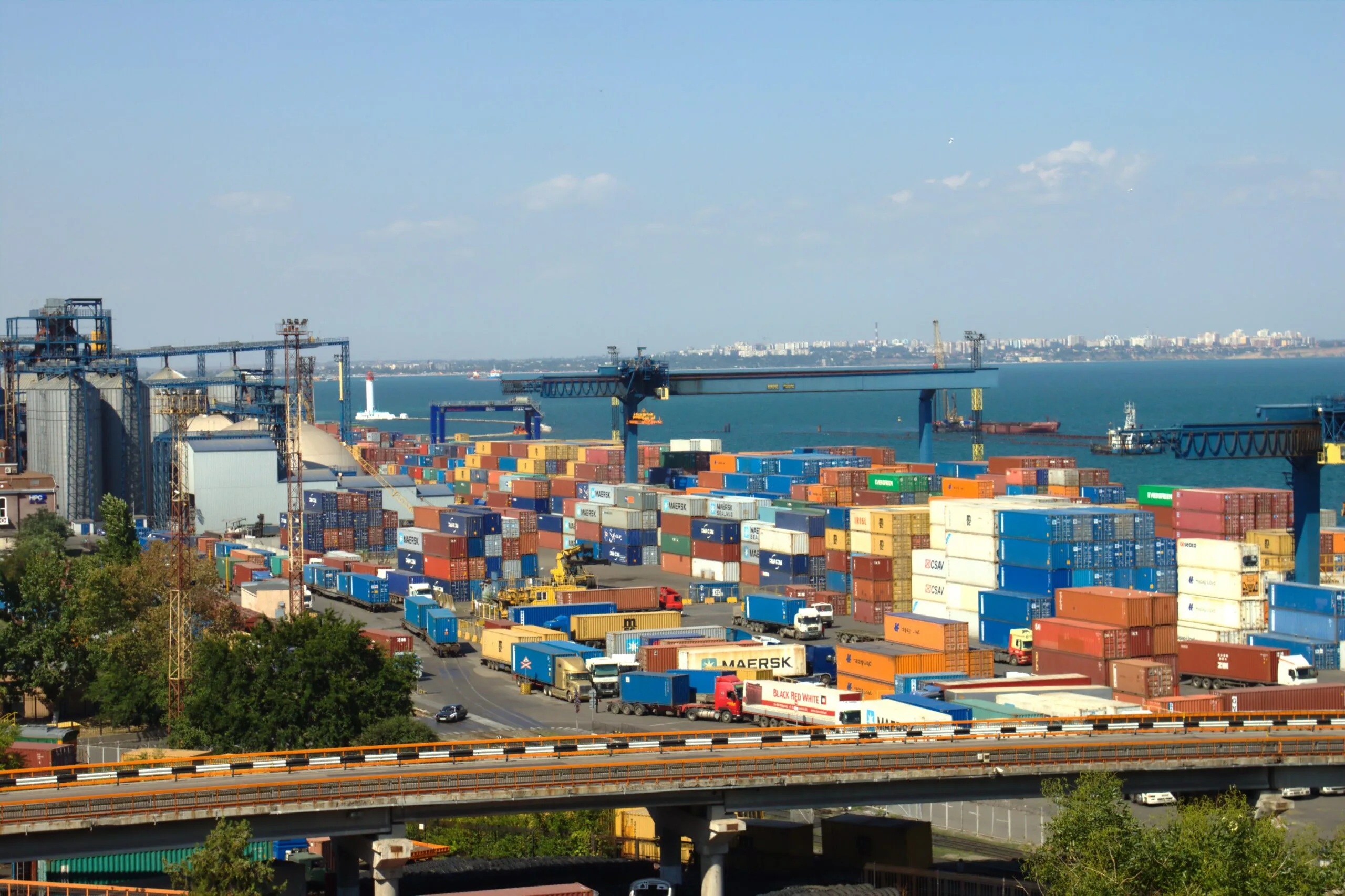Terminale portuale con centinaia di container colorati impilati, gru di carico e navi ancorate in acque blu.