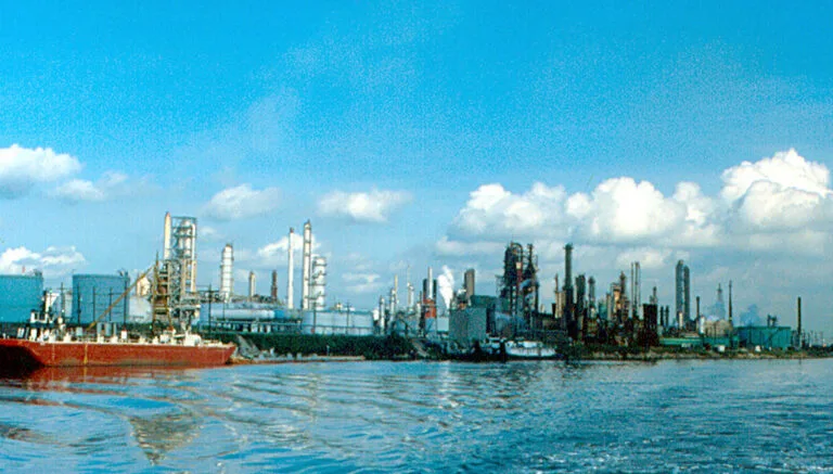 Complesso industriale petrolifero con torri di raffinazione e una nave cisterna rossa in primo piano su acque blu.