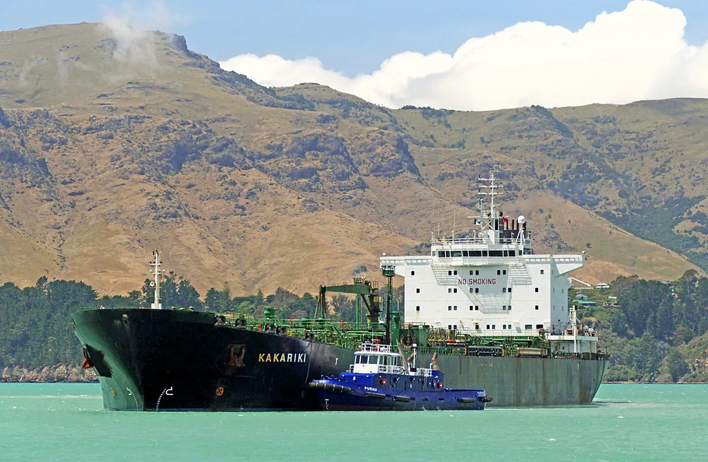 Una petroliera nera con il nome KAKARIKI affiancata da un rimorchiatore blu in acque turchesi davanti a colline aride.
