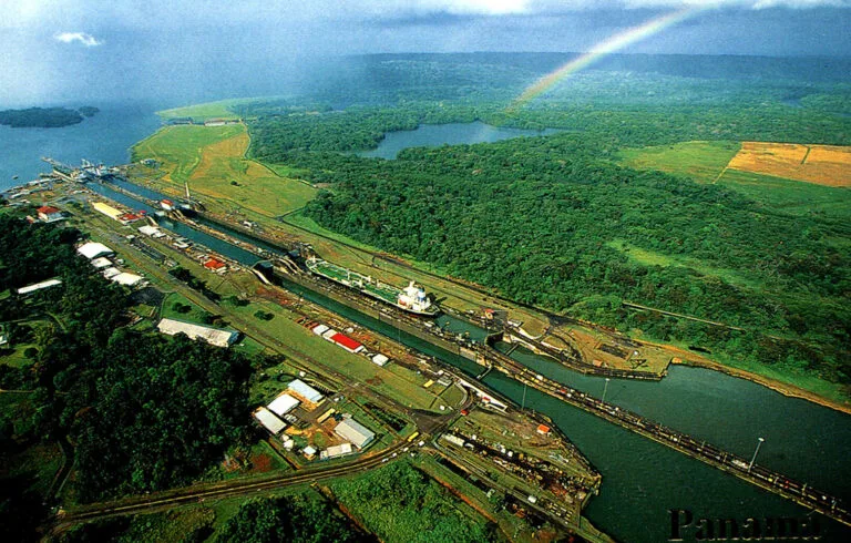 Veduta aerea del Canale di Panama con navi portacontainer in transito, circondato da foresta tropicale e zone industriali.