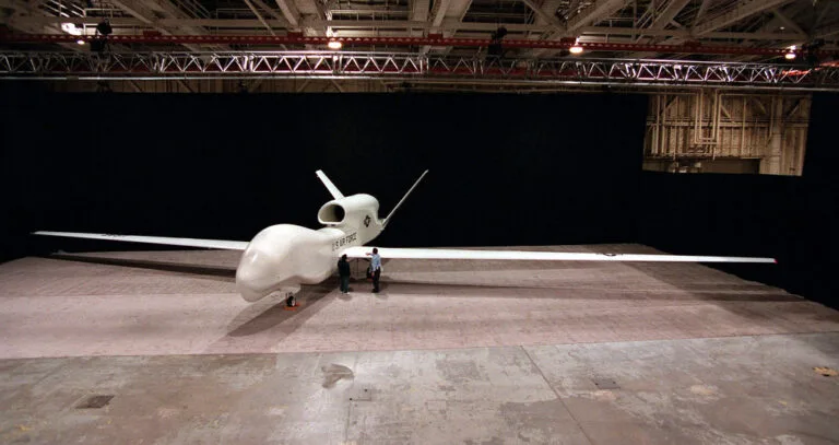 Drone tattico bianco con ali lunghe e fusoliera arrotondata esposto in un hangar industriale illuminato.
