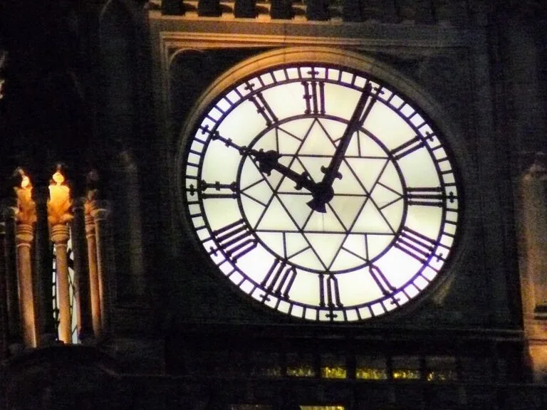 Grande orologio illuminato con numeri romani su una torre buia, fiaccole accese ai lati.