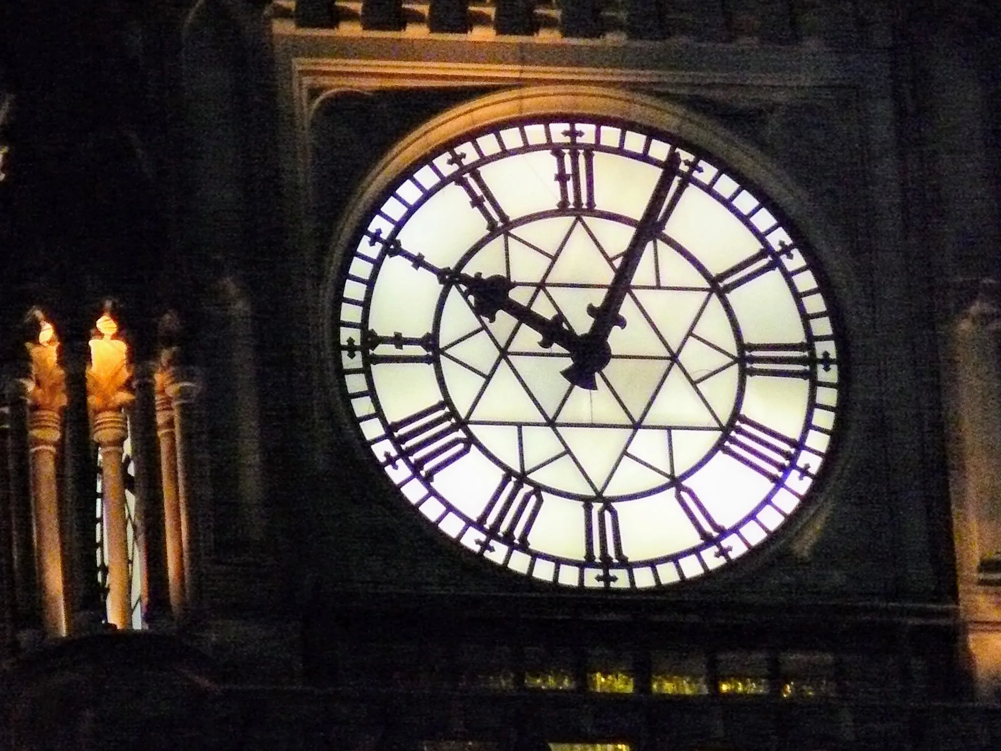 Grande orologio illuminato con numeri romani su una torre buia, fiaccole accese ai lati.