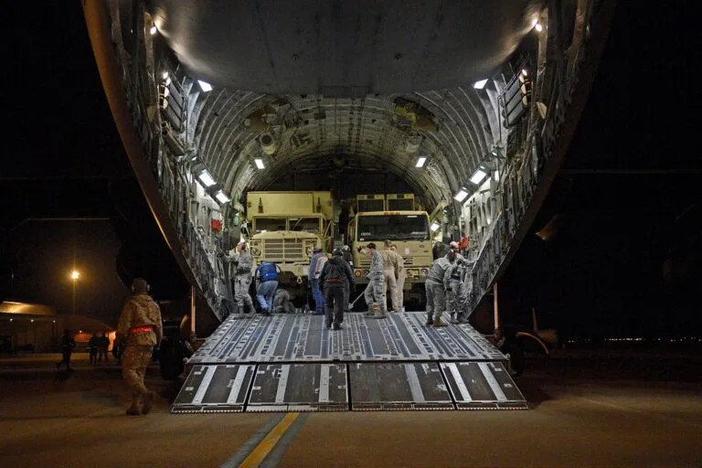 Aereo militare cargo con portellone aperto di notte scarica veicoli blindati su una pista.