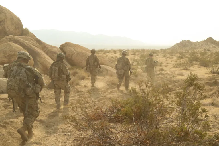 Soldati in uniforme percorrono un terreno desertico arido tra formazioni rocciose e vegetazione rada.