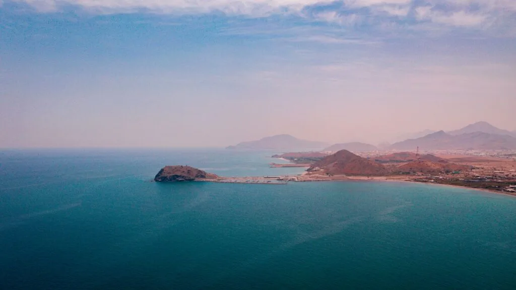 Costa desertica con spiaggia sabbiosa, isola rocciosa e catene montuose sullo sfondo affacciate su acque blu tranquille.