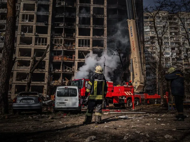 Vigili del fuoco intervengono su un edificio residenziale danneggiato con fiamme e fumo denso mentre soldati osservano.