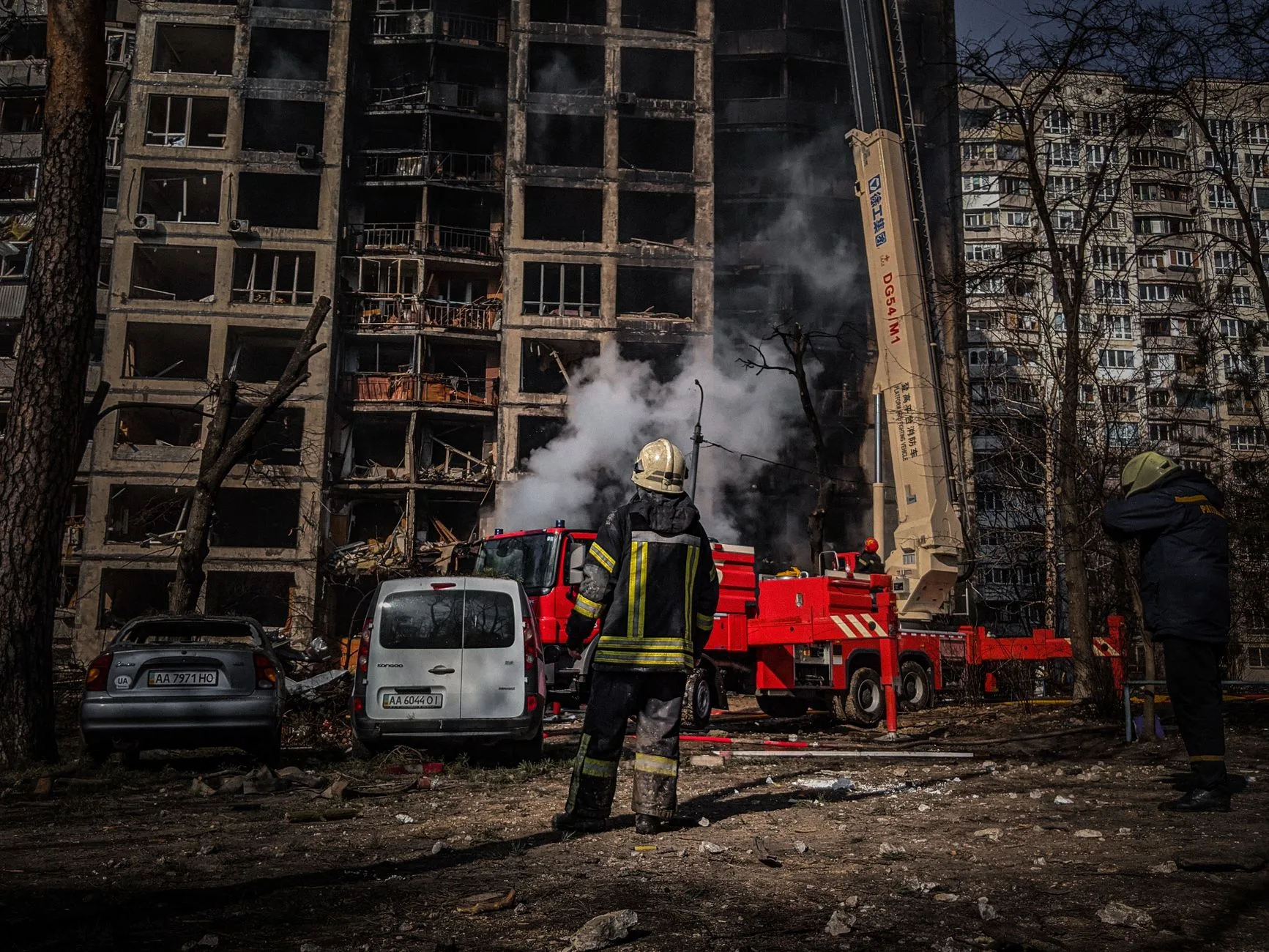 Vigili del fuoco intervengono su un edificio residenziale danneggiato con fiamme e fumo denso mentre soldati osservano.