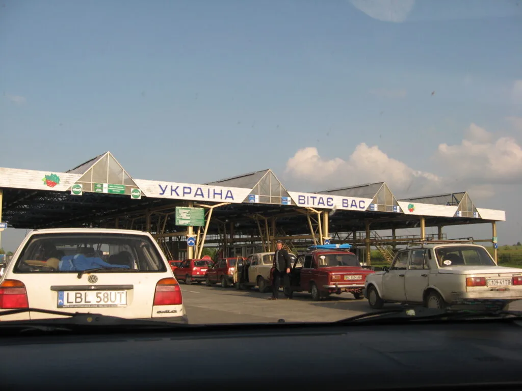 Auto in fila a un checkpoint di frontiera con insegna in cirillico che riporta «Ucraina» e «Vitae Vas», cielo sereno.