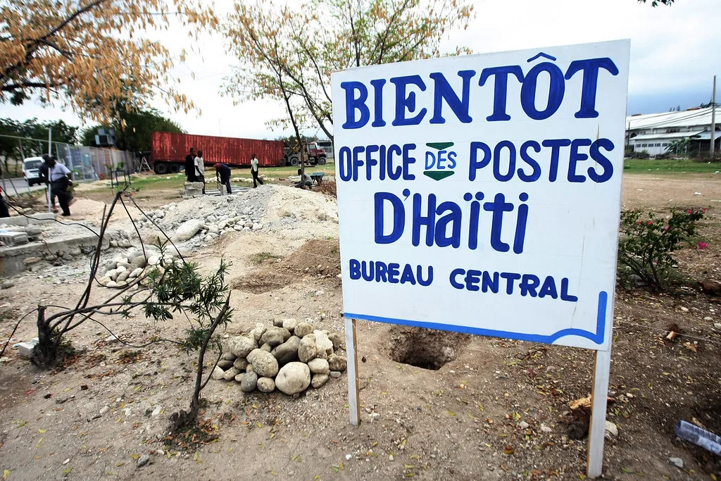 Cartello dell'ufficio postale centrale di Haiti in un'area con edifici rovinati e persone sullo sfondo.
