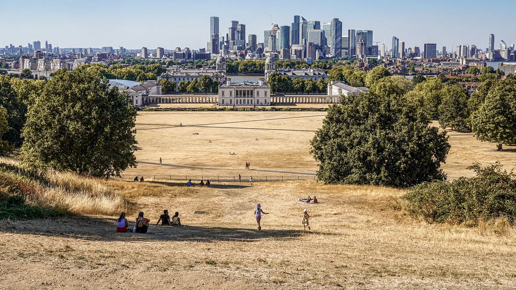 Parco pubblico con erba secca, persone sedute all'ombra, edificio storico e skyline di città sullo sfondo.