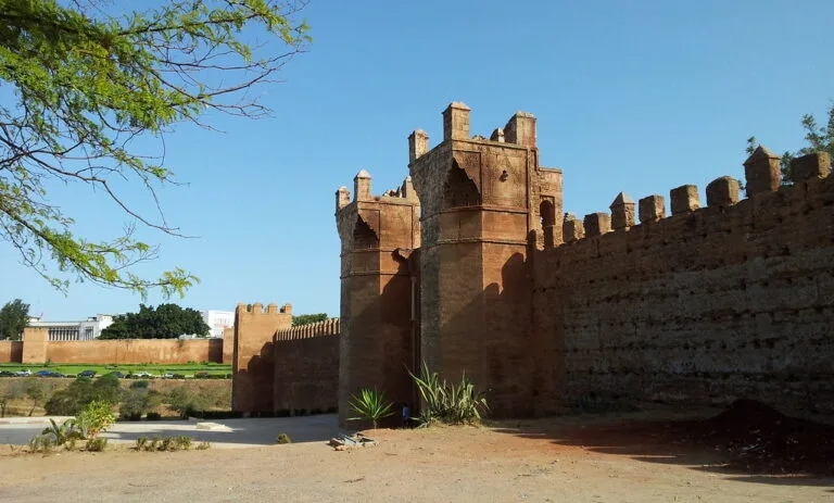 Fortificazione in mattoni rossi con torri merlate e porte monumentali, circondata da mura massicce sotto cielo sereno.