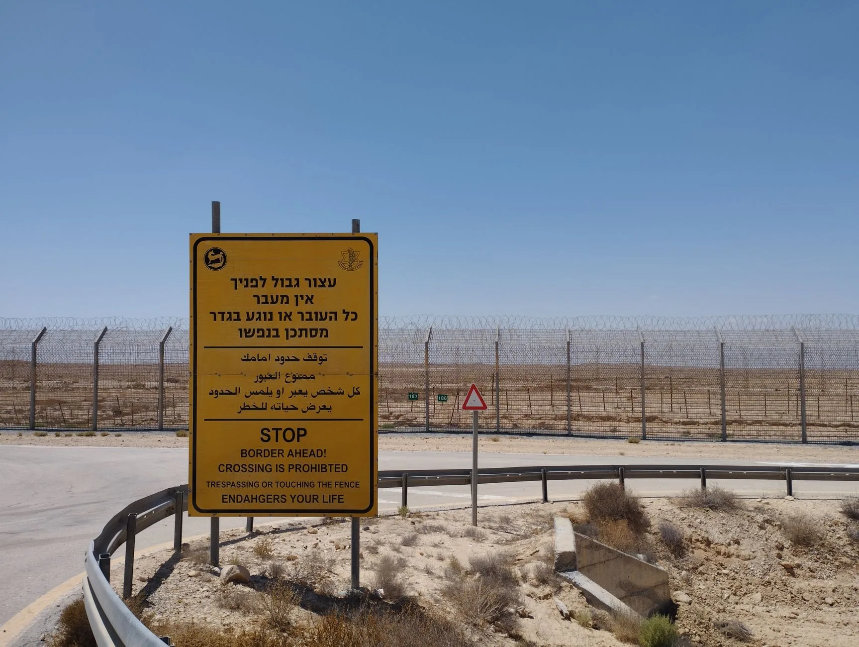 Cartello di avvertimento giallo in ebraico e inglese davanti a recinzione metallica in zona desertica al confine.
