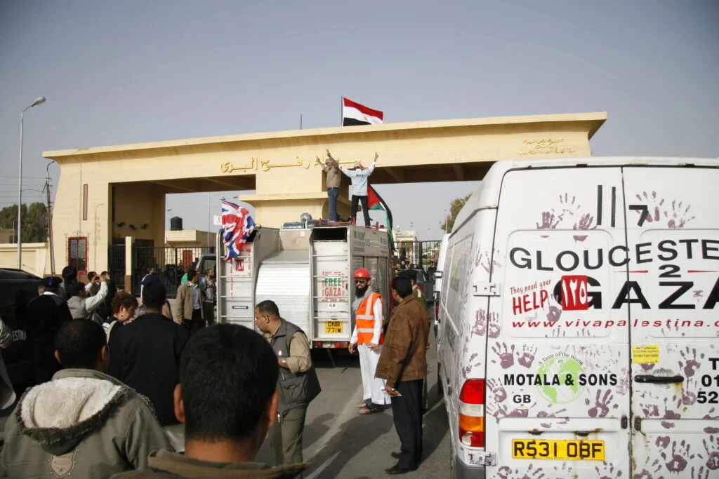Varchi di frontiera al Rafah Crossing con bandiera egiziana, veicoli di aiuti umanitari e persone in attesa.