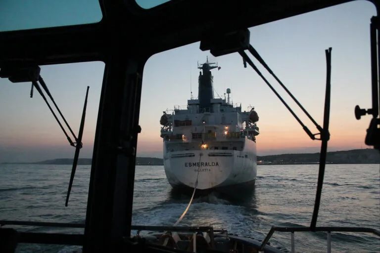 Una nave cargo fotografata al tramonto da un'altra imbarcazione, con il nome Esmeralda visibile sulla prua illuminata.