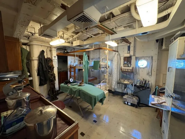 Interno di una nave ospedale con letti a castello, attrezzature mediche e banchi di lavoro lungo le pareti metalliche.
