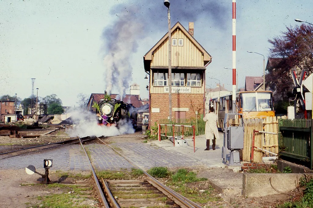 Locomotiva a vapore in stazione ferroviaria con edificio residenziale in mattoni e camino industriale sullo sfondo.