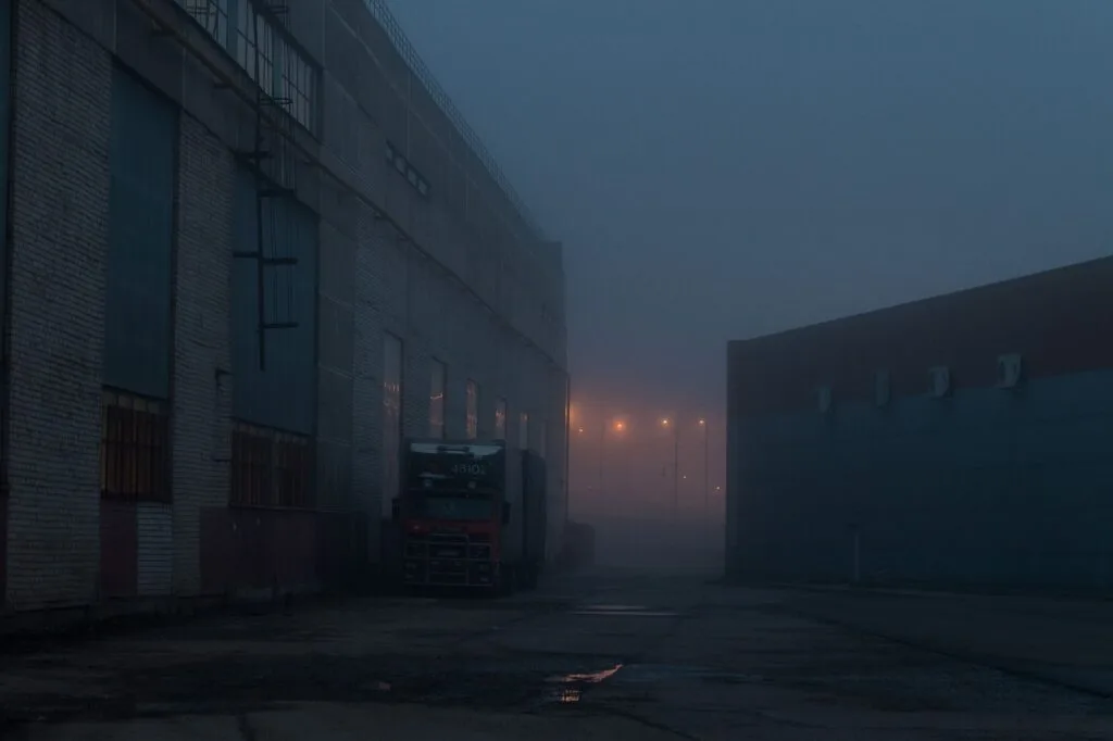 Magazzino industriale avvolto dalla nebbia di notte, con camion parcheggiato e luci stradali che brillano nel buio.