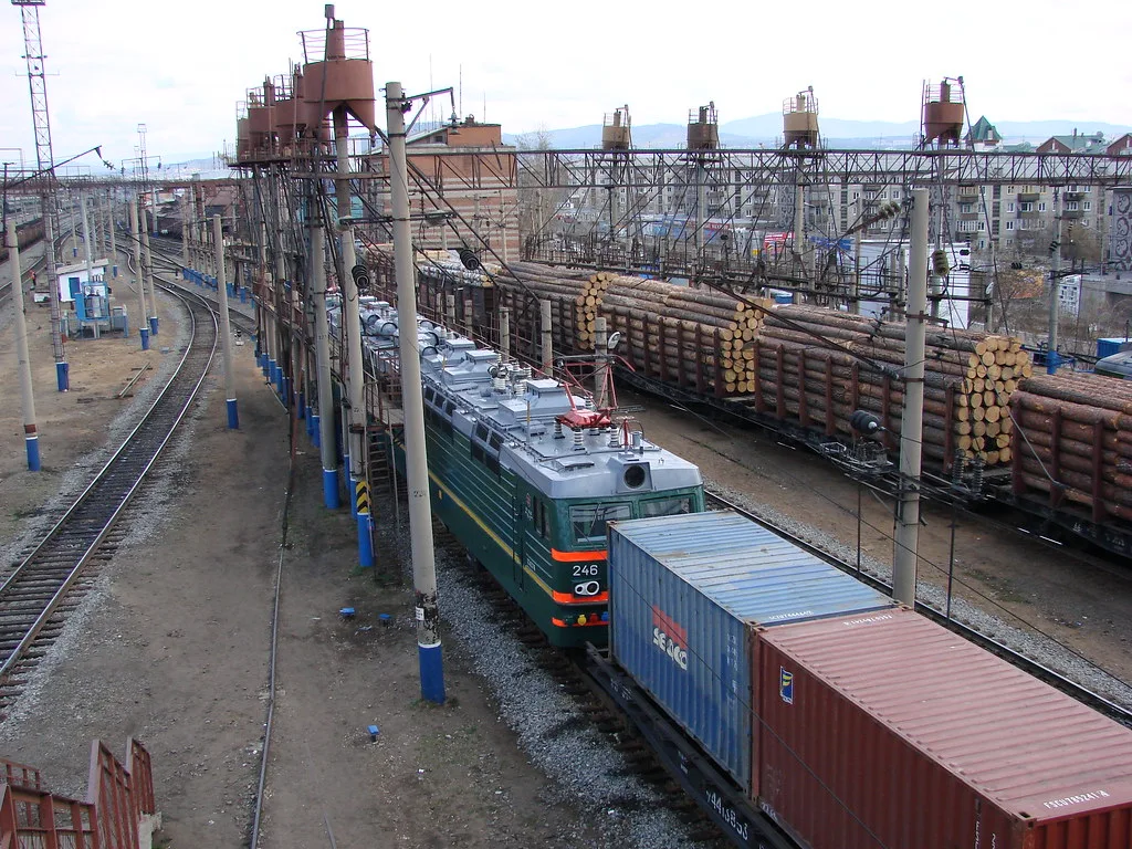 Stazione ferroviaria con locomotiva verde e vagoni merci, infrastrutture di carico e cumuli di tronchi d'albero sullo sfondo.
