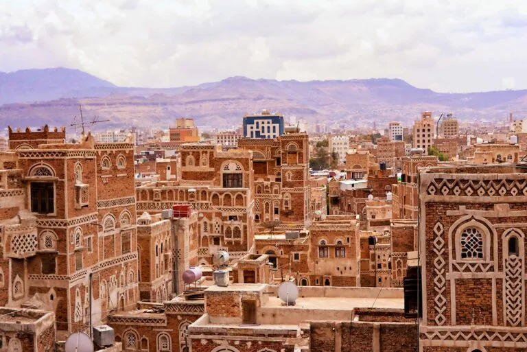 Veduta aerea della città vecchia di Sanaa con edifici in laterizio decorato e montagne sullo sfondo.