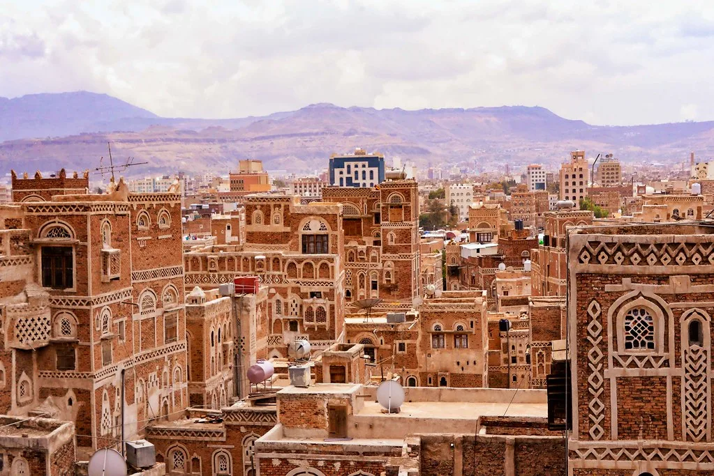 Veduta aerea della città vecchia di Sanaa con edifici in laterizio decorato e montagne sullo sfondo.