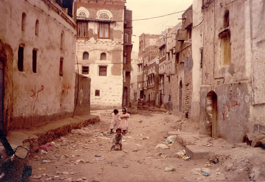 Bambini giocano in una strada polverosa tra edifici danneggiati e fatiscenti.