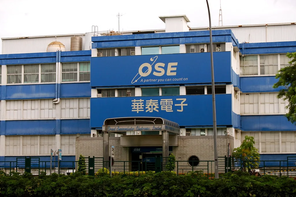 Facciata di un edificio industriale con strisce blu e bianche, insegna OSE e caratteri cinesi, circondato da vegetazione.