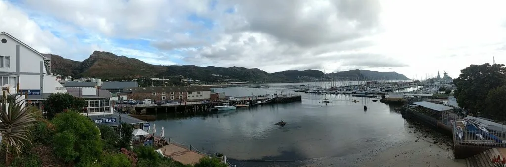 Porto di Simon's Town con barche ormeggiate, banchine e montagni sullo sfondo sotto cielo nuvoloso.
