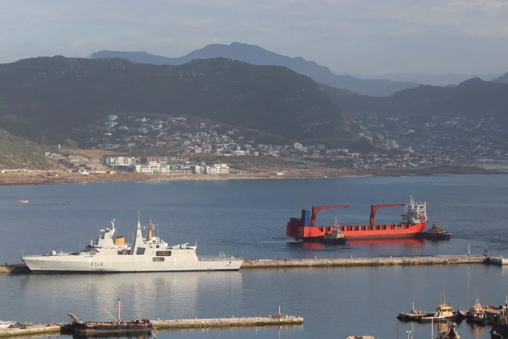 Navi militari e commerciali ormeggiate in un porto naturale circondato da colline e insediamenti costieri.