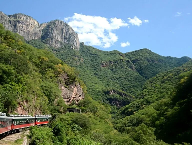 Treno rosso che percorre una vallata montagnosa rigogliosa con pareti rocciose a strapiombo.