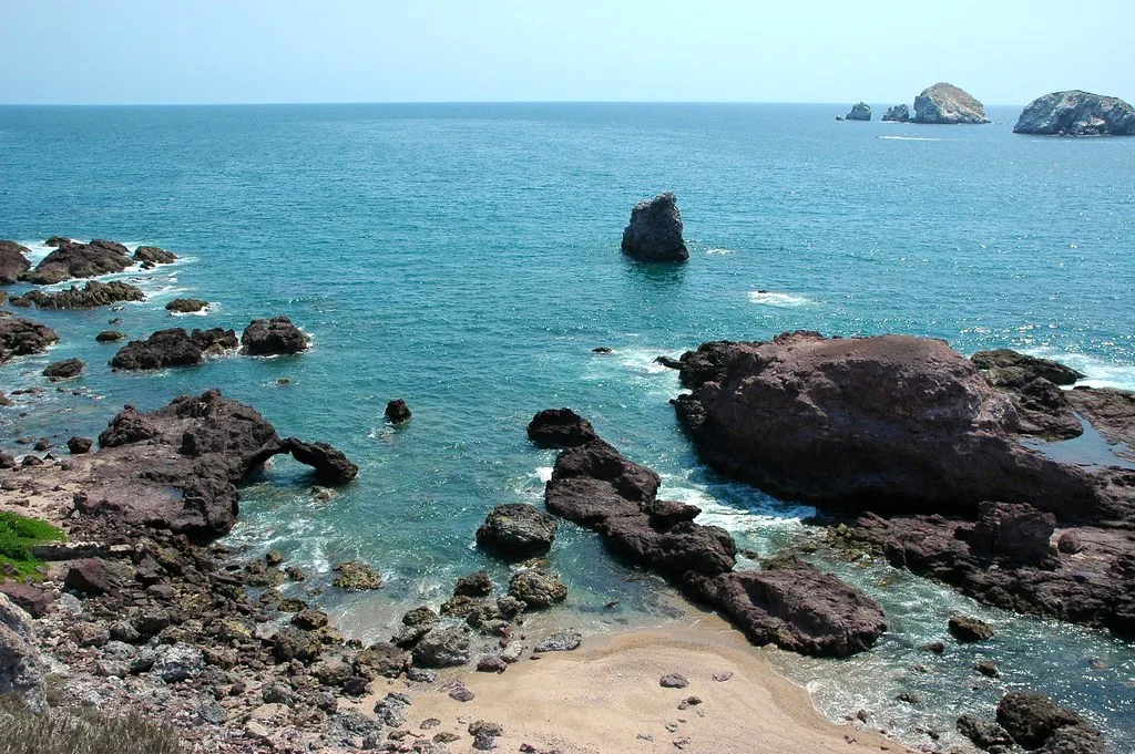 Costa rocciosa con spiaggia sabbiosa, formazioni basaltiche e acque turchesi del Mediterraneo.