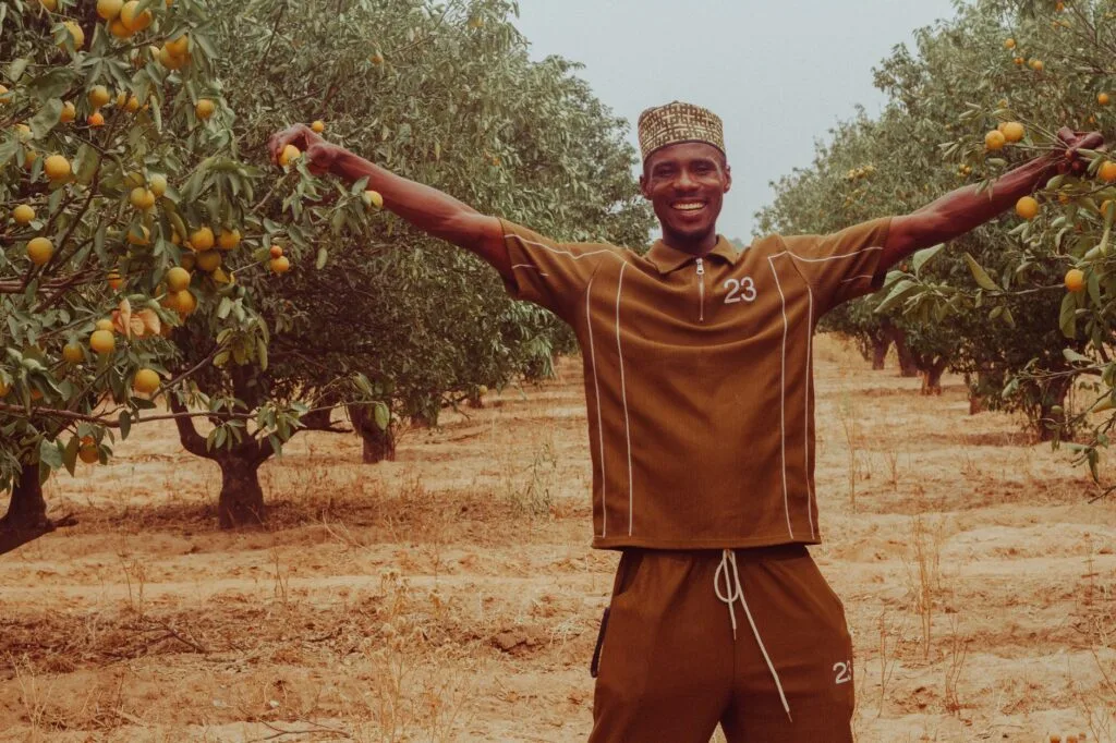 Uomo sorridente con le braccia aperte in un frutteto di agrumi, circondato da alberi carichi di frutti.