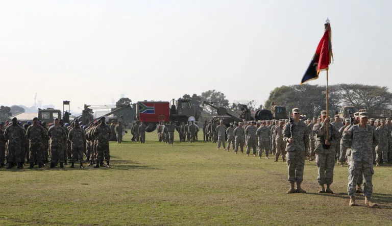 Truppe in uniforme schierati in formazione su un prato con bandiera sudafricana durante cerimonia militare.