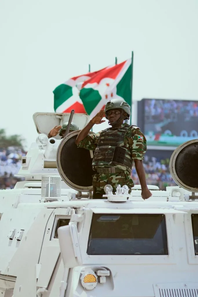 Soldato in uniforme mimetica saluta da veicolo blindato bianco durante parata militare con bandiera tricolore.